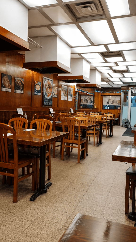 Cozy restaurant interior featuring traditional warm wood paneled walls, wooden tables, chairs, and framed Korean artwork under bright ceiling panels.