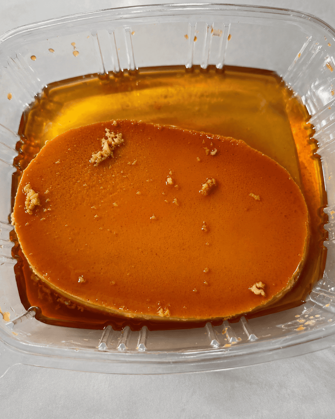 A smooth, oval-shaped Filipino Leche Flan custard sitting in sweet, golden caramel syrup inside a clear plastic takeout container.