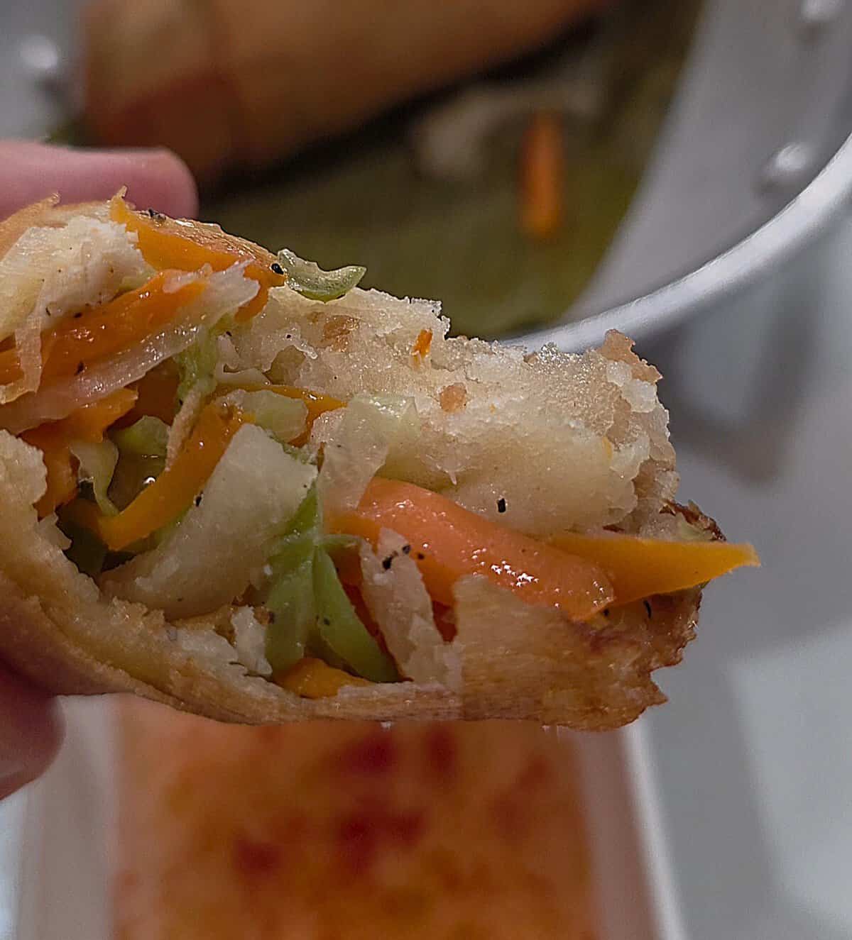 A close-up hand-held shot of a crispy Filipino fried lumpia prito bitten in half, showing a savory filling of shredded carrots, cabbage, and chicken.