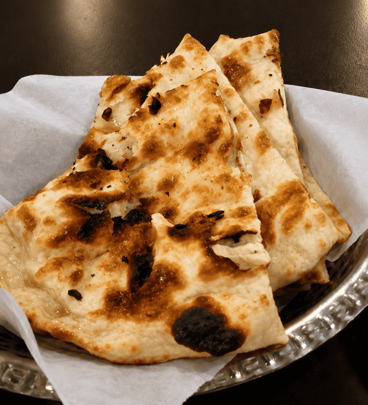 Slices of charred, blistered, and buttery naan bread stacked neatly inside a metal basket lined with white parchment paper.