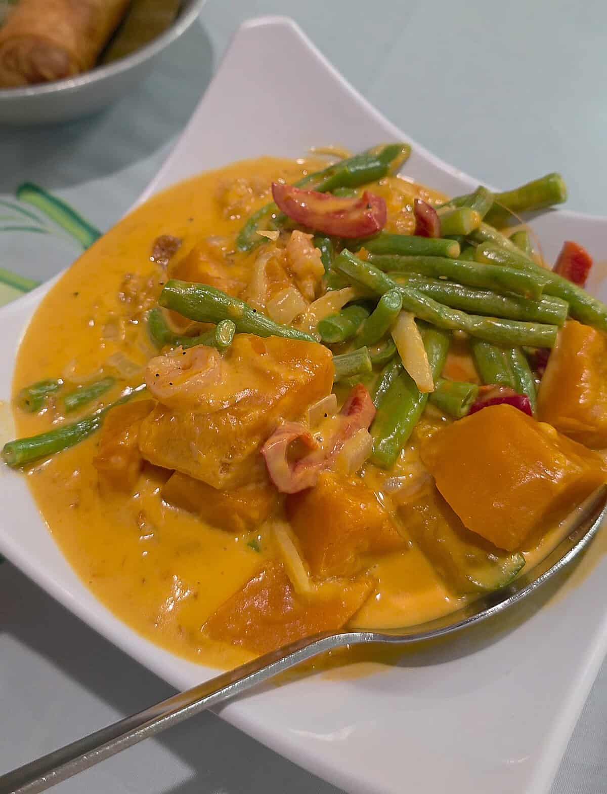 A bowl of Ginataang Kalabasa with Sitaw, featuring chunks of yellow squash, bright green yardlong beans, and shrimp simmered in a creamy coconut milk sauce.