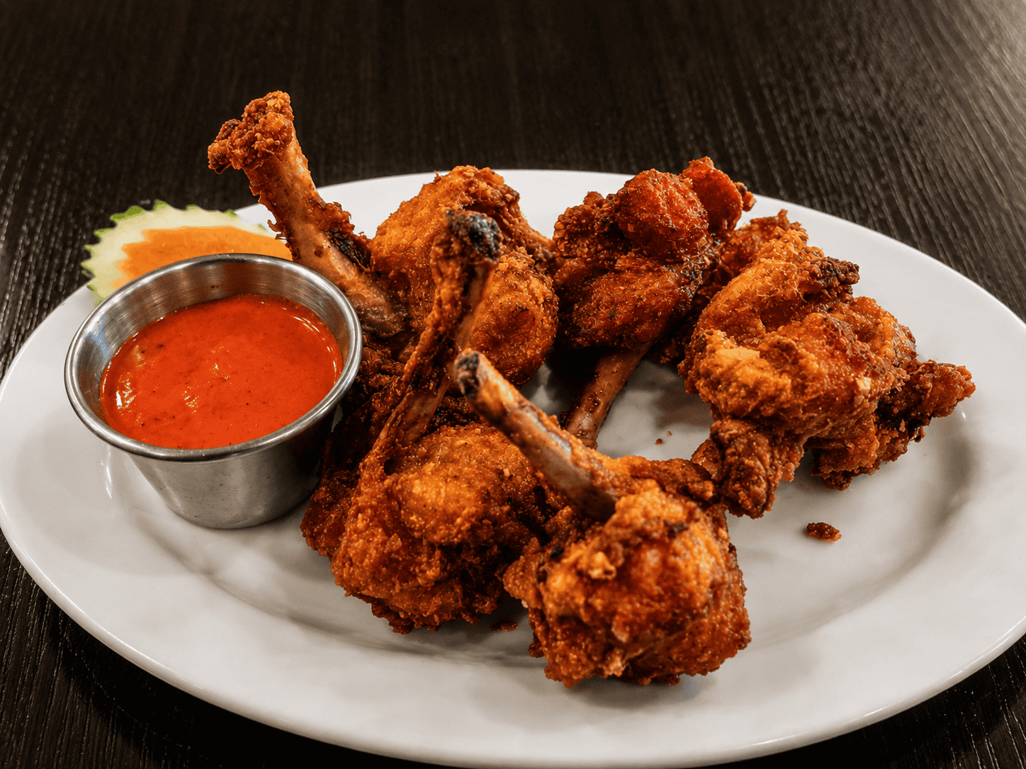 A white plate holding a pile of deep-fried Chicken Lollipops with exposed bone handles, served next to a small metal cup filled with bright red dipping sauce.