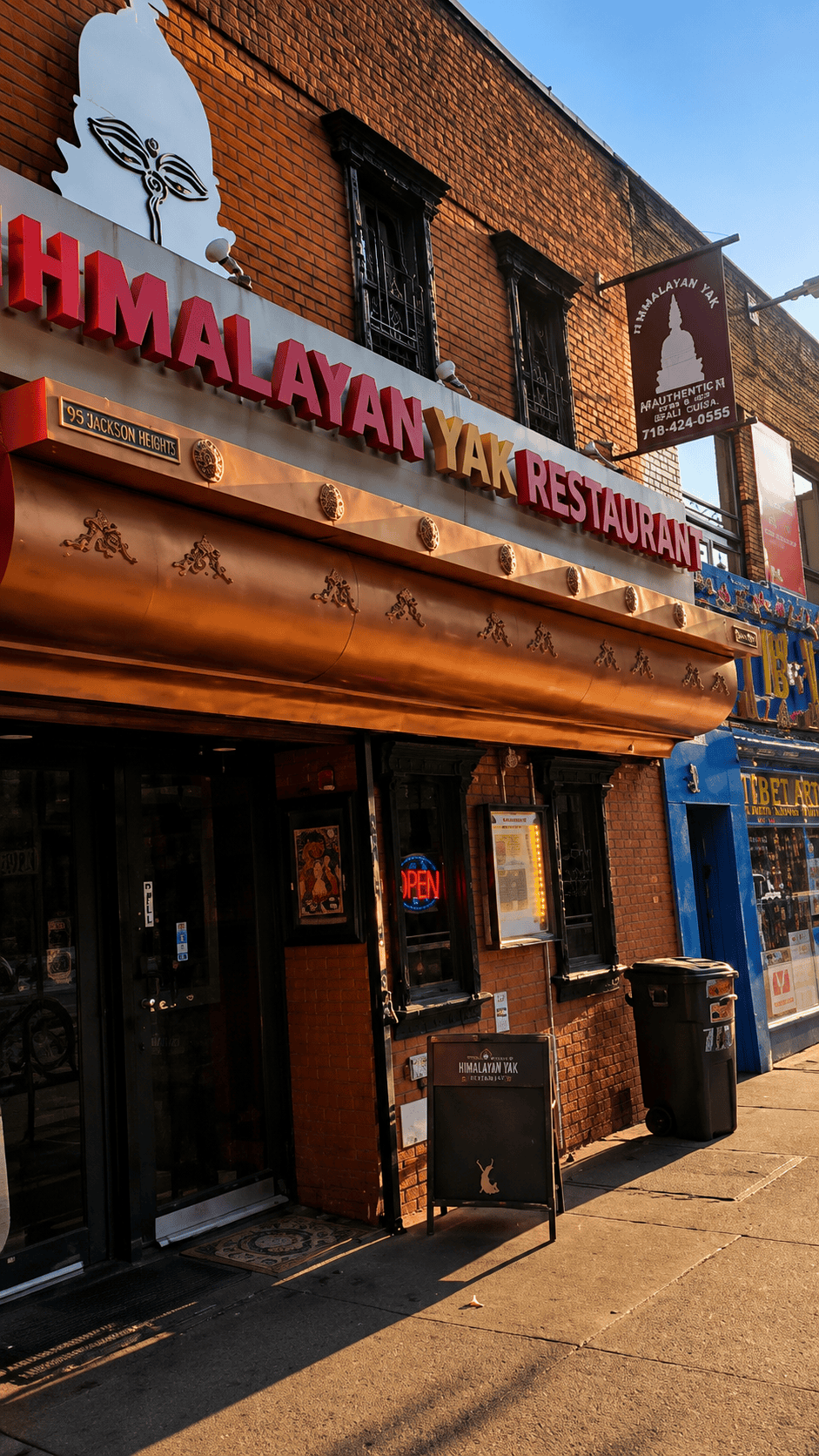 The brick exterior and storefront of Himalayan Yak Restaurant in Jackson Heights featuring a prominent copper-colored awning, neon open sign, and a large white traditional stupa eye emblem above the restaurant name.
