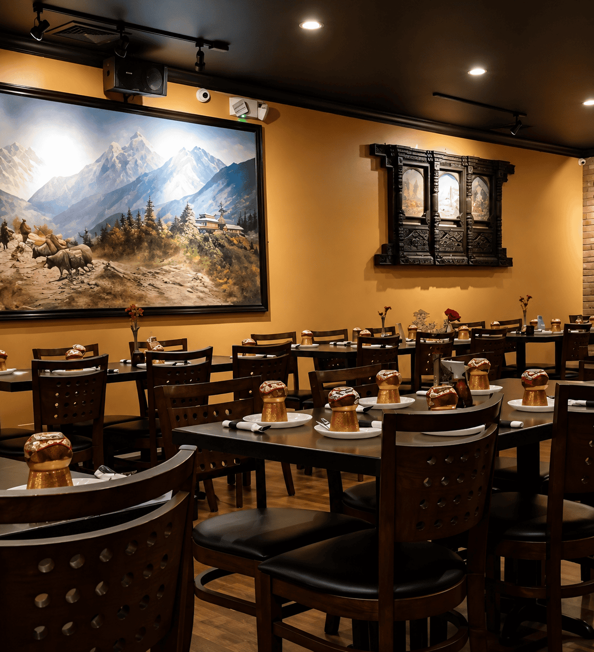 The modern, clean interior dining room of Himalayan Yak featuring dark wood tables and chairs, yellow walls, a large painting of Himalayan mountains and yaks, and traditional wooden window frames.