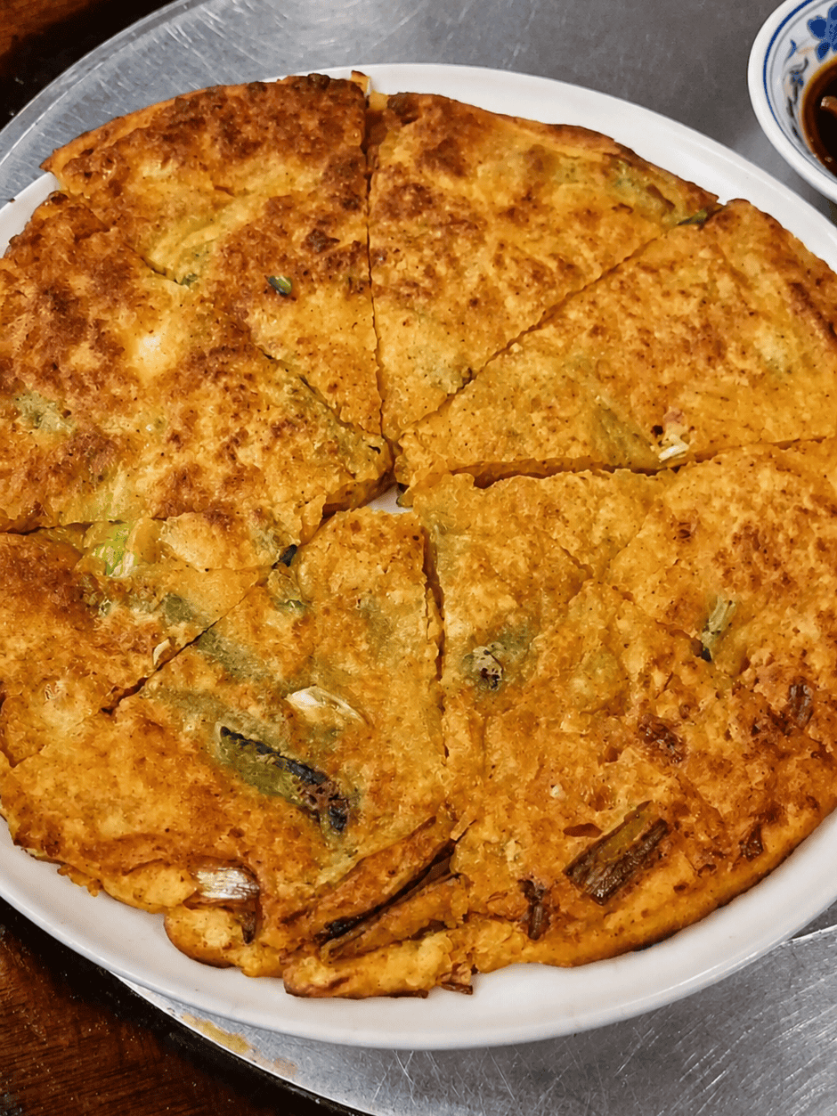 An orange-red crispy Kimchi Pajun savory Korean pancake sliced into wedges on a white plate next to a small dish of dipping sauce.