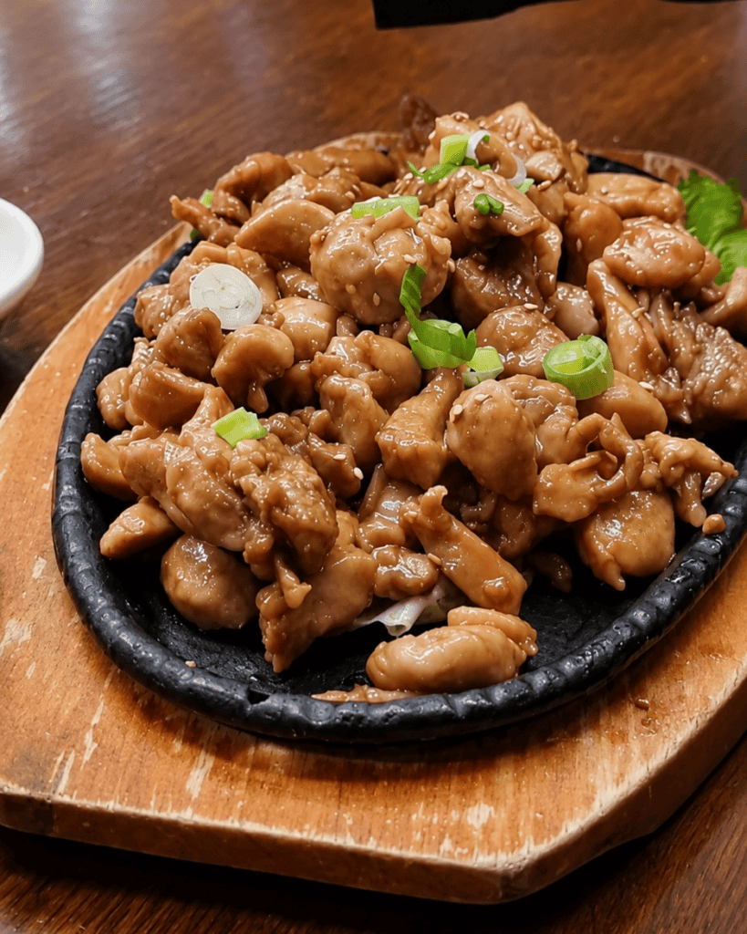 Sizzling cast iron plate on a wooden board filled with Korean style grilled chicken breast pieces seasoned with sesame seeds and garnished with chopped green scallions.