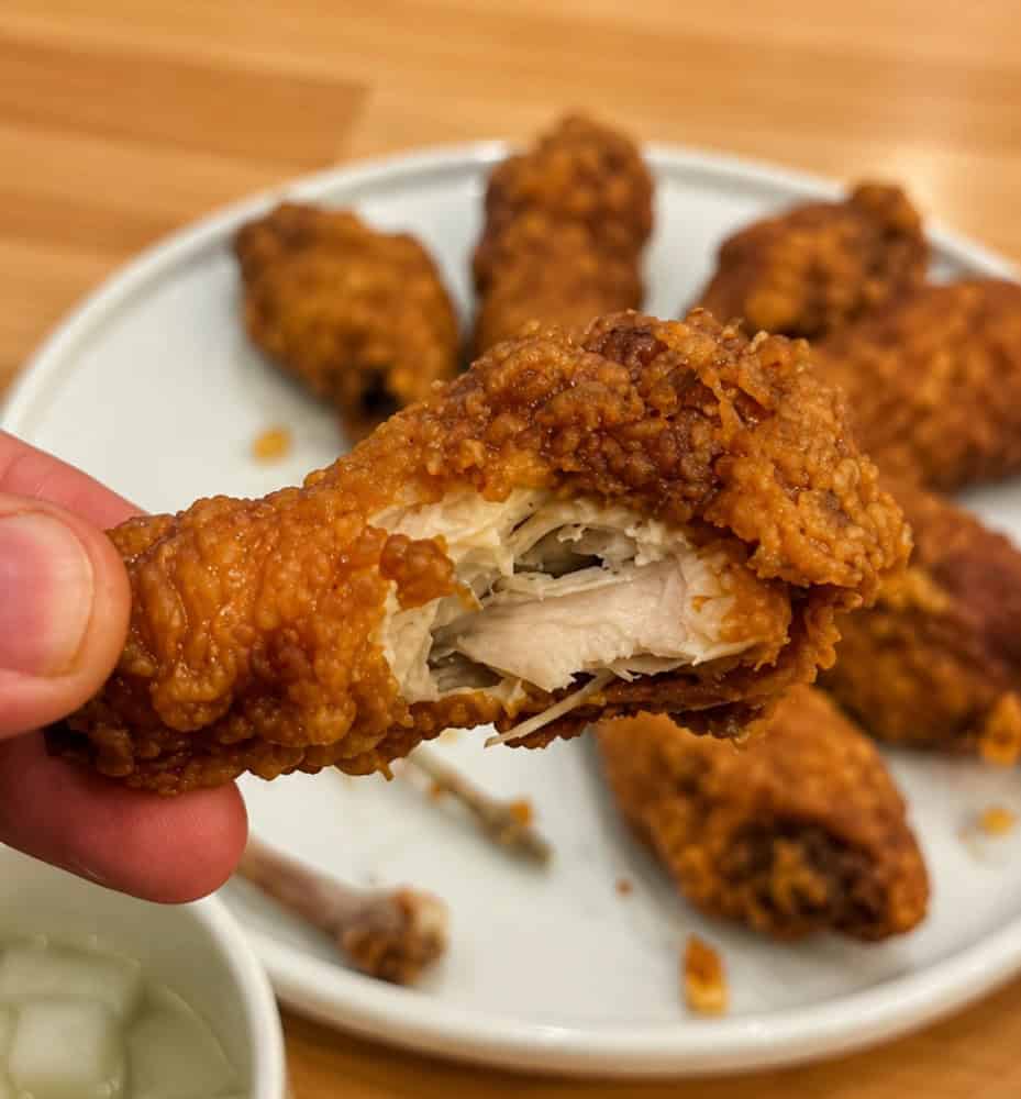 A hand holding a crispy soy garlic Korean fried chicken wing with a bite taken out, showing the juicy meat inside.