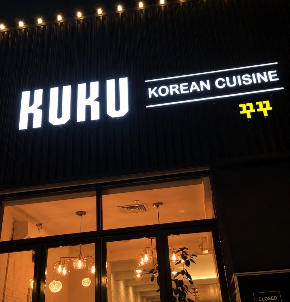 Exterior storefront of Kuku Korean Cuisine in Forest Hills at night, showing a modern dark facade and large glowing sign on 70th Road.