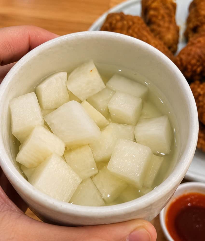 A close-up view of a small white bowl filled with cubed Korean pickled radish in a clear brine.