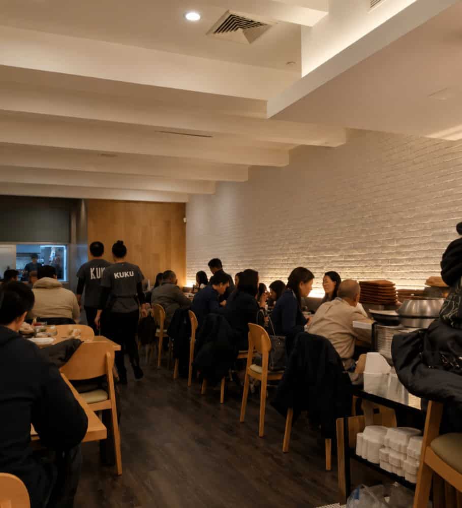 Interior view of Kuku's modern dining room with contemporary lighting, sleek white brick accents, and tables filled with dining customers.