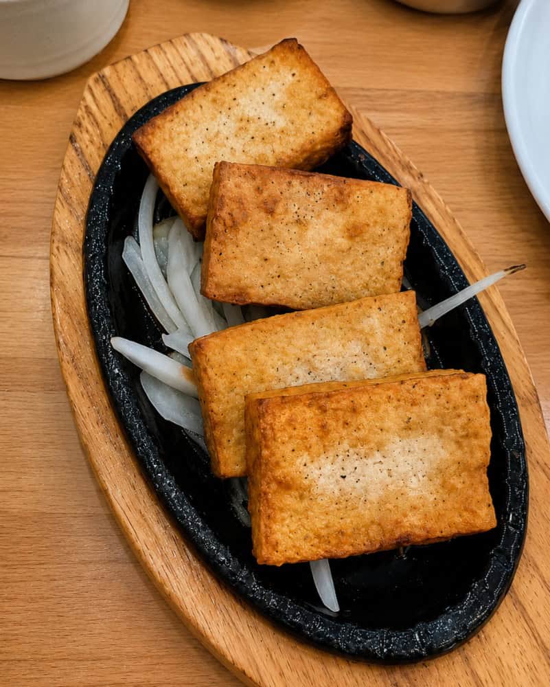 Golden-brown, perfectly crisped rectangular blocks of fried tofu served sizzling over a small bed of sliced white onions in a cast iron dish.