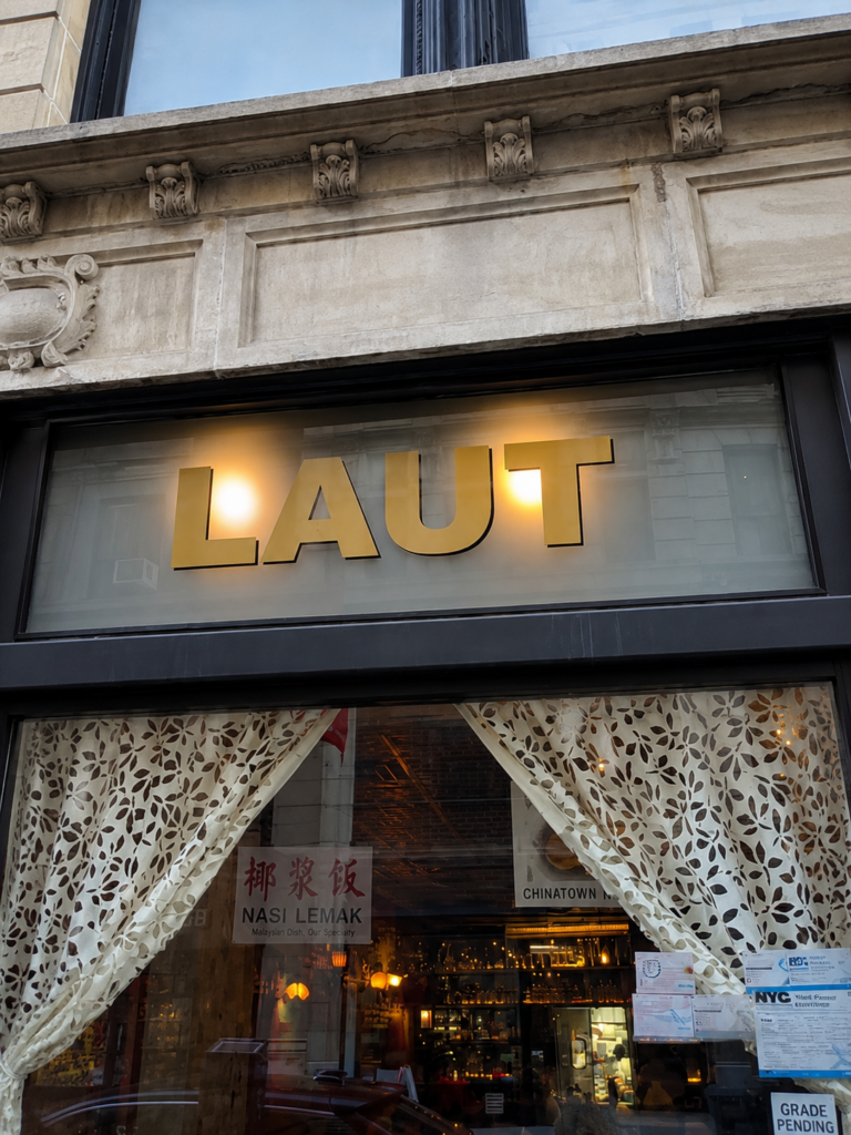 The gold illuminated exterior sign of Laut restaurant in Manhattan with patterned curtains in the window