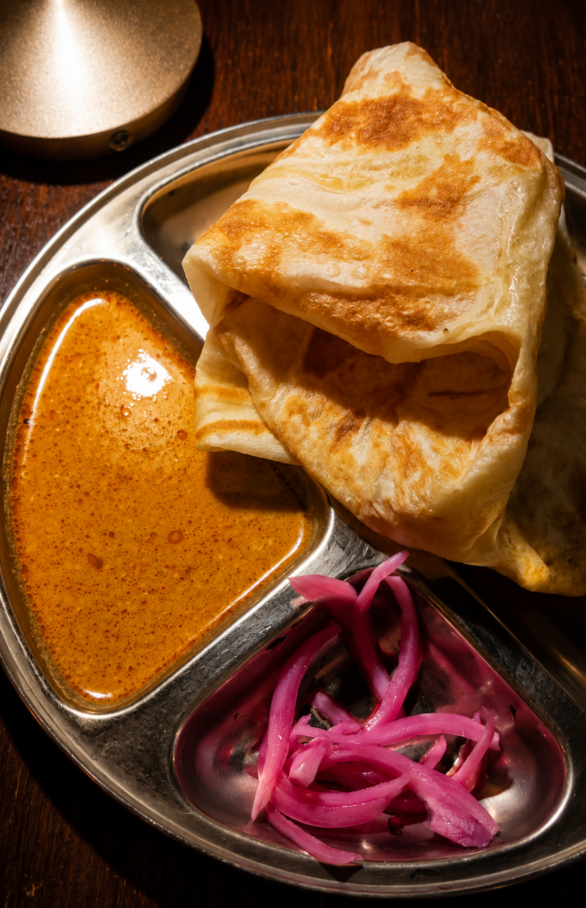 Golden flaky Malaysian roti canai served on a metal platter with curry dipping sauce and pickled red onions