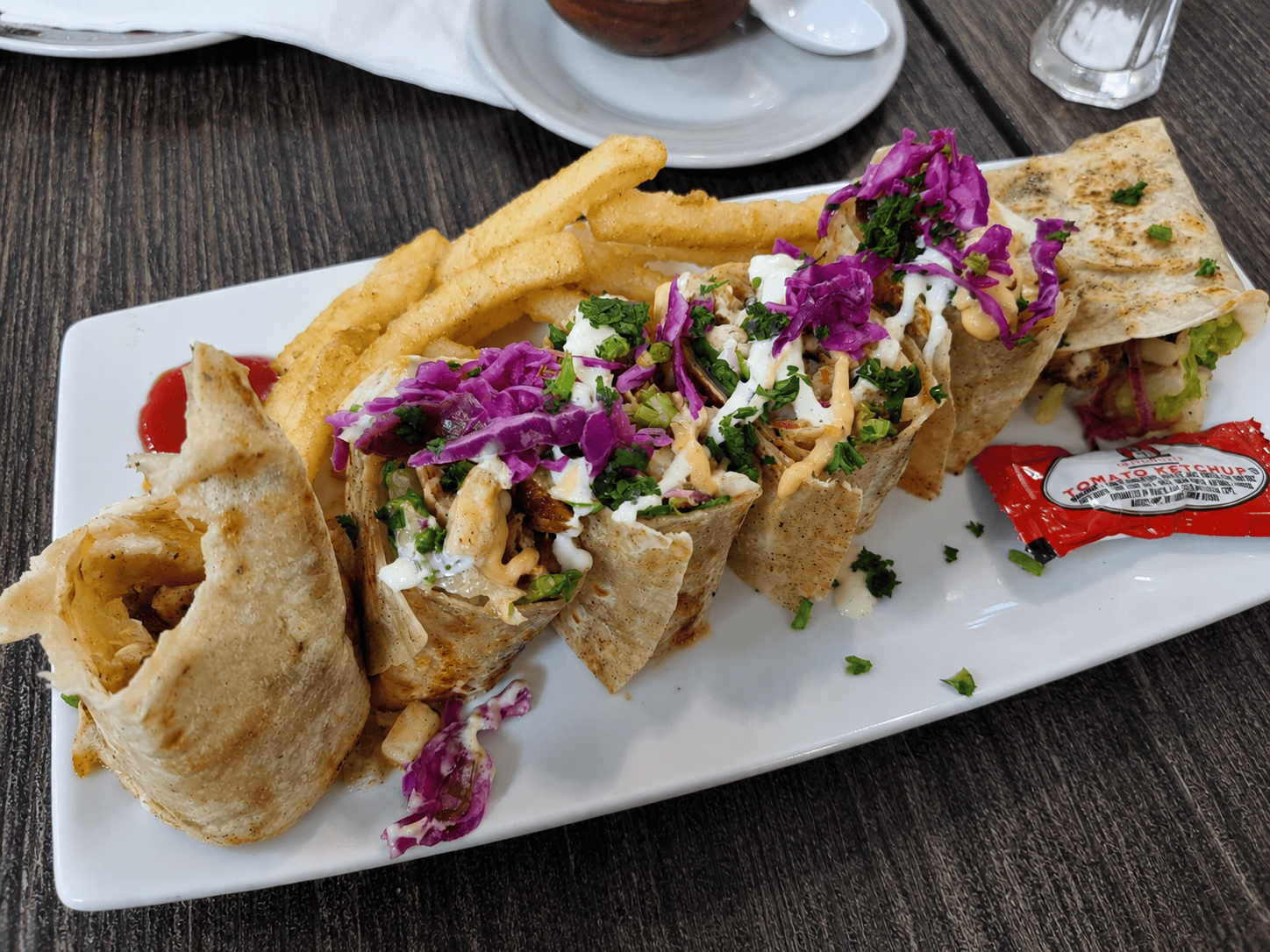 A chicken shawarma wrap neatly sliced into sections on a white plate, topped with shredded red cabbage and herbs, served with golden french fries and a ketchup packet.