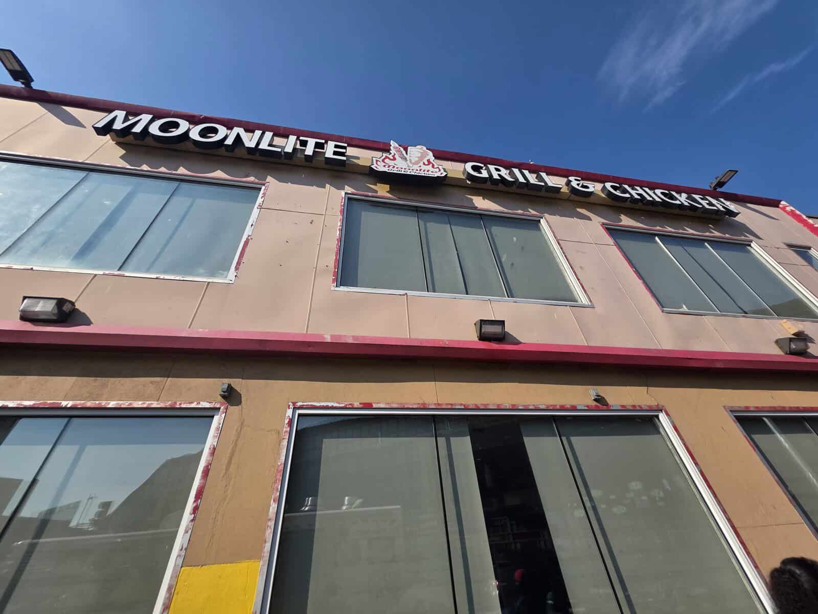 The exterior storefront of Moonlite Grill and Chicken in Jackson Heights, Queens, showcasing its pink and tan facade against a bright blue sky.