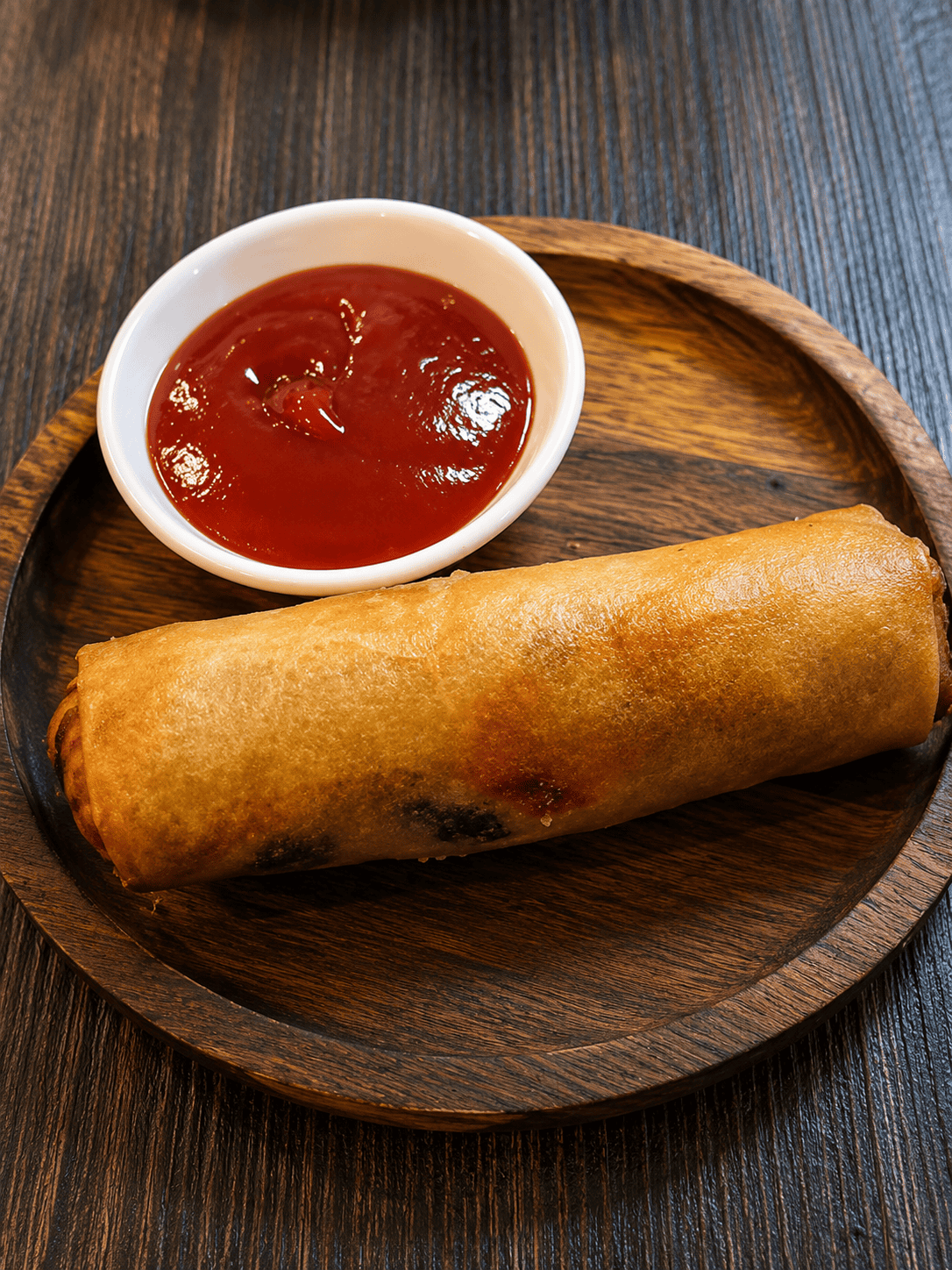 A golden-brown, crispy fried chicken roll served on a round wooden plate next to a small white bowl of red ketchup.