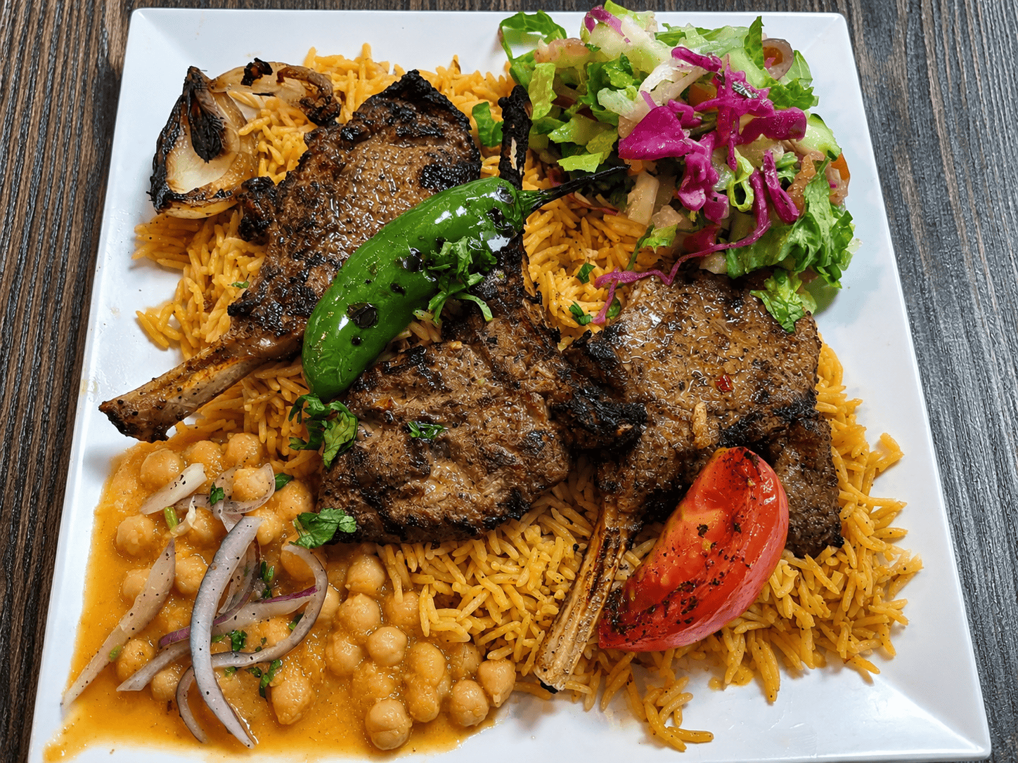 A heavily loaded platter of grilled lamb chops over orange seasoned rice, served with chickpeas, a charred green pepper, a grilled tomato, and a red cabbage side salad.