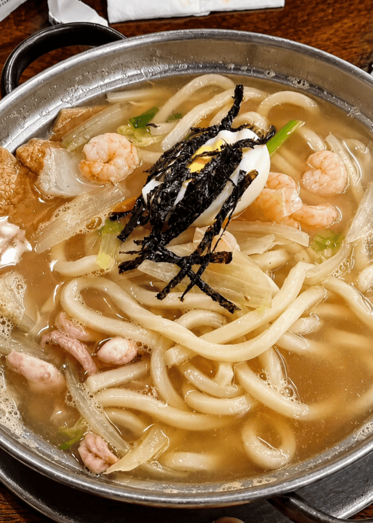 Nam Bi Udon noodle soup served in a traditional metal pot filled with thick wheat noodles, plump shrimp, vegetables, shredded seaweed, and a boiled egg slice.