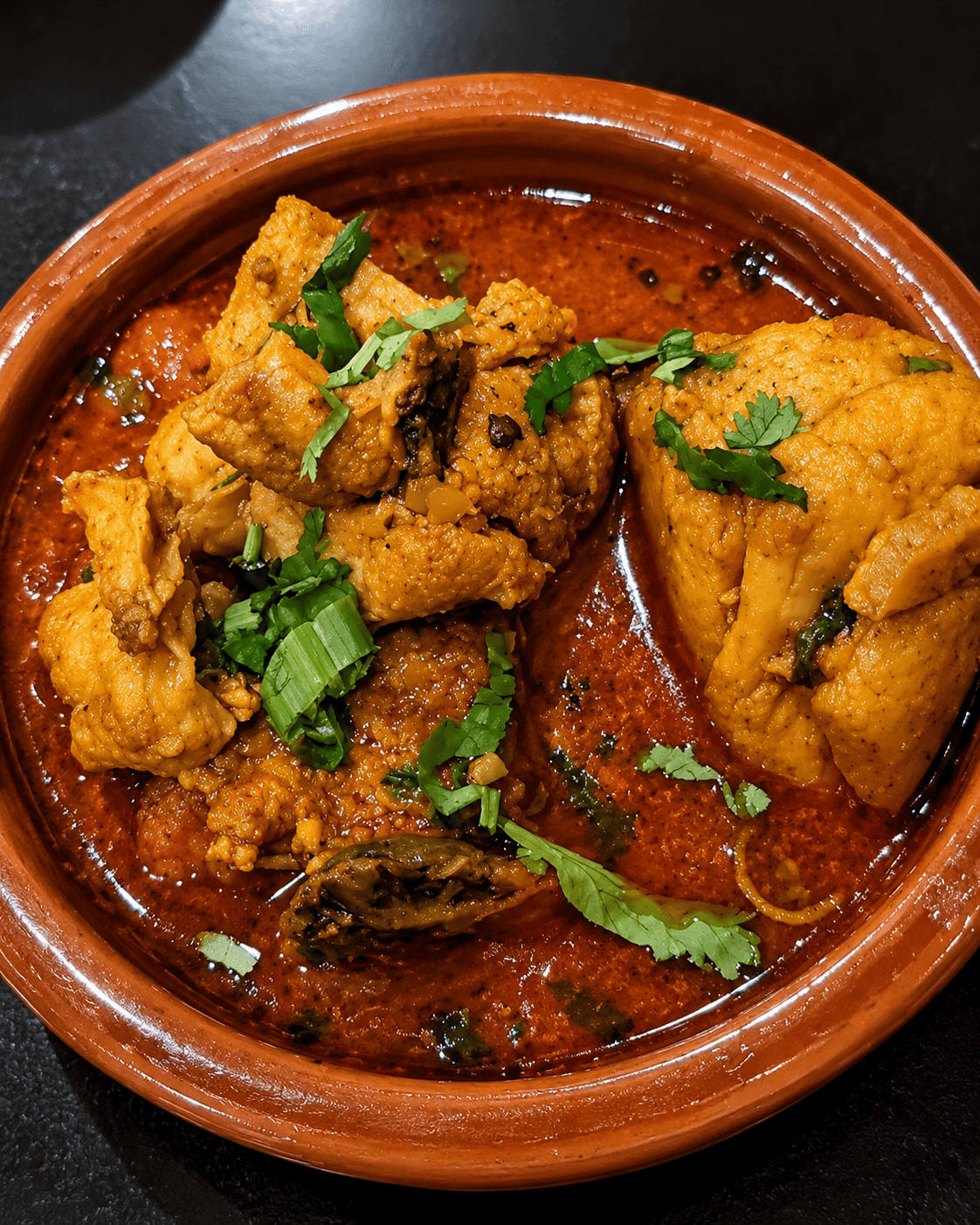 A rich orange-red chicken curry garnished with fresh green cilantro and scallions, served inside a rustic brown earthenware clay dish.