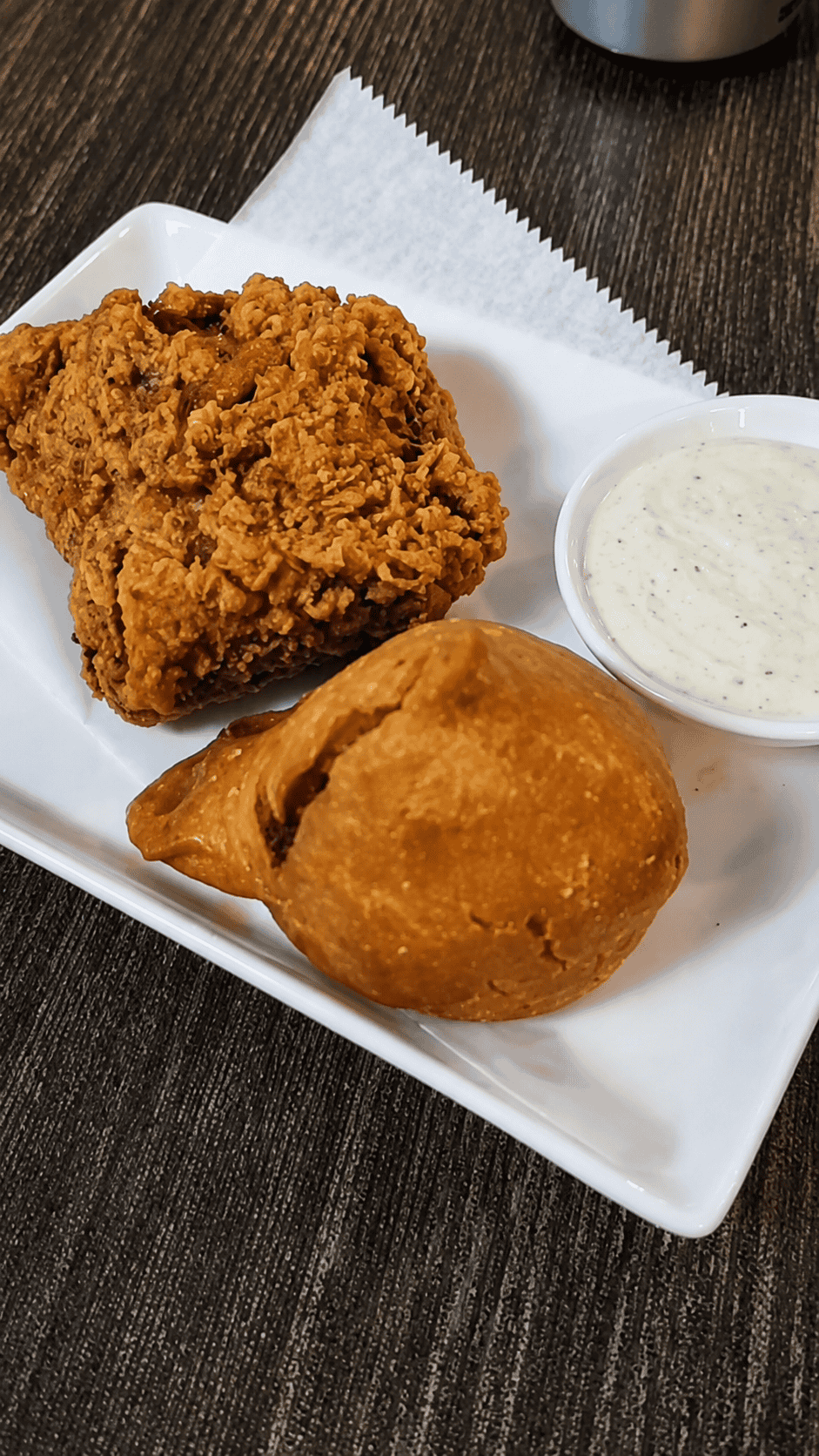 A white plate holding a piece of crispy, golden-brown fried chicken, a fried samosa, and a small side dish of white garlic yogurt sauce.