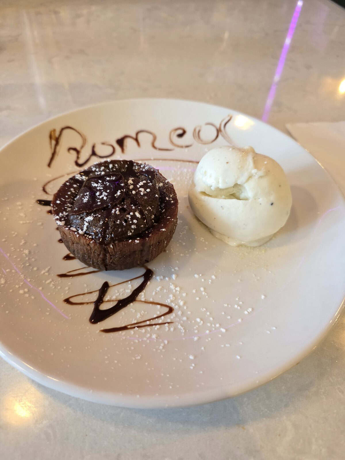 A dessert plate featuring a round, warm chocolate soufflé dusted with powdered sugar alongside a scoop of vanilla ice cream, with 'Romeo' written elegantly in chocolate drizzle.