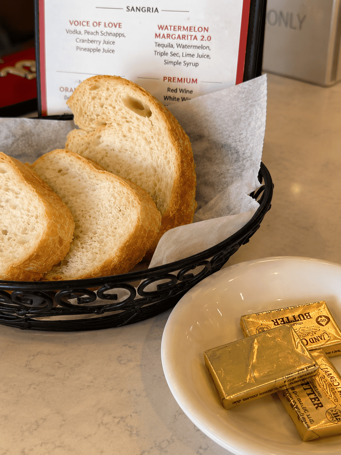 A black wire basket containing thick slices of crusty Italian white bread served with gold foil-wrapped Land O Lakes butter pats on a small white dish.