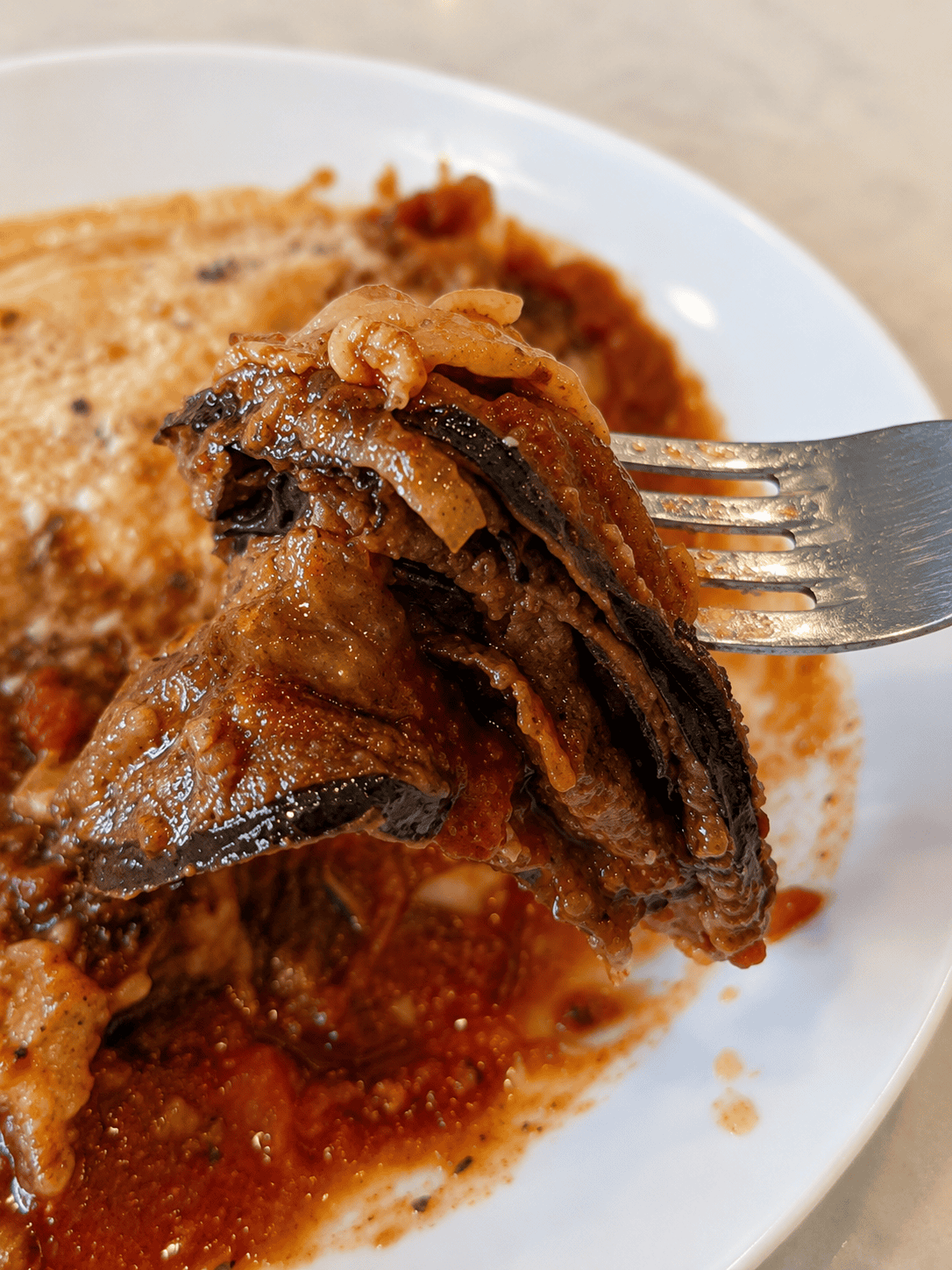 A close-up view of a metal fork lifting a layered bite of tender, soft baked eggplant coated in rich marinara sauce.