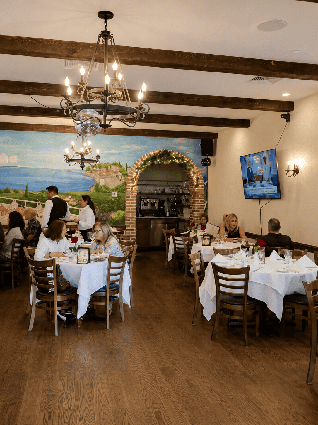 Elegant dining room interior of Romeo's Restaurant with white tablecloths, wooden chairs, exposed ceiling beams, a large chandelier, and a colorful painted mural on the back wall.