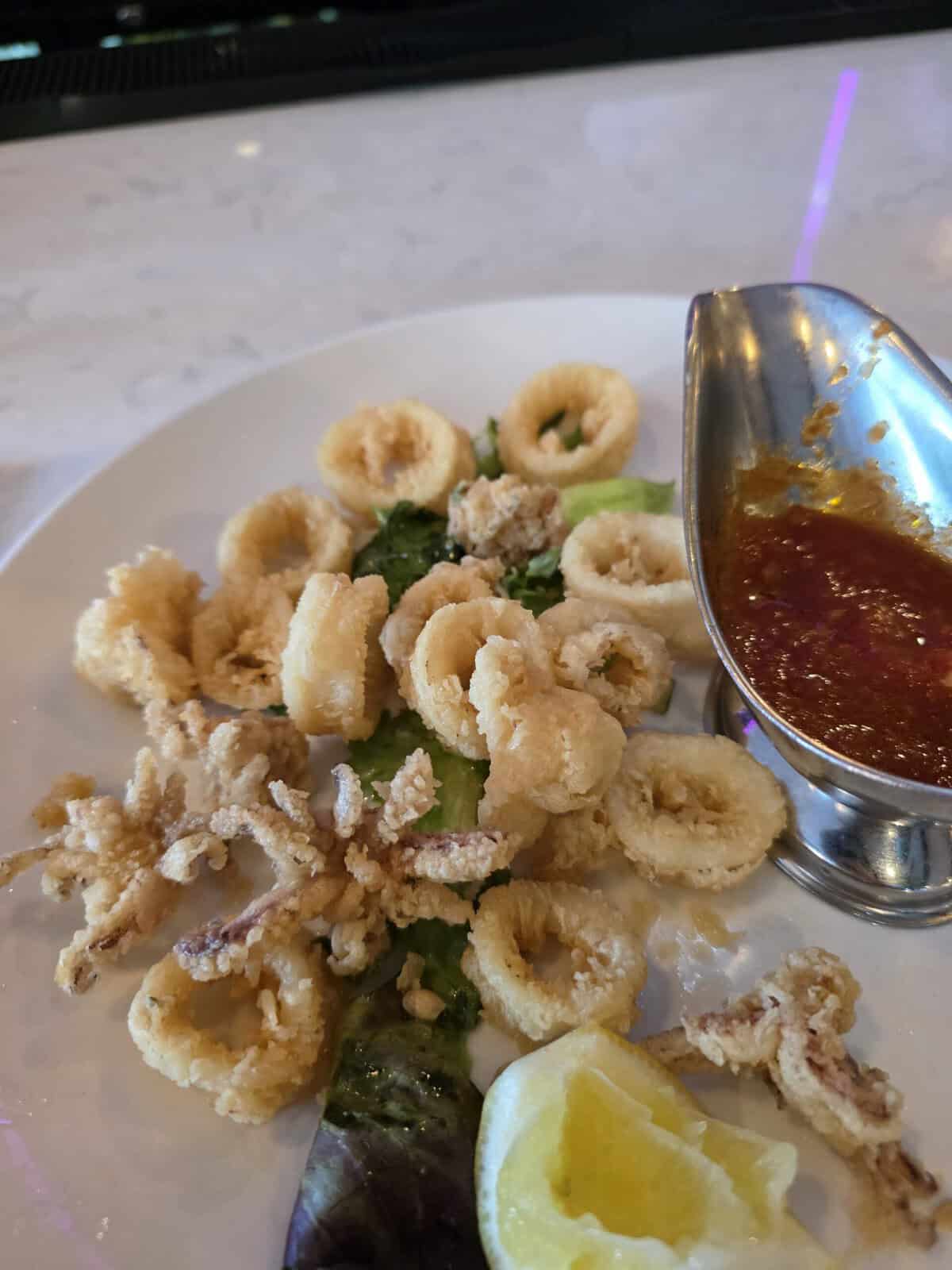 A white plate piled high with golden, crispy fried calamari rings and tentacles, served with a lemon wedge and a metal boat of red marinara sauce.