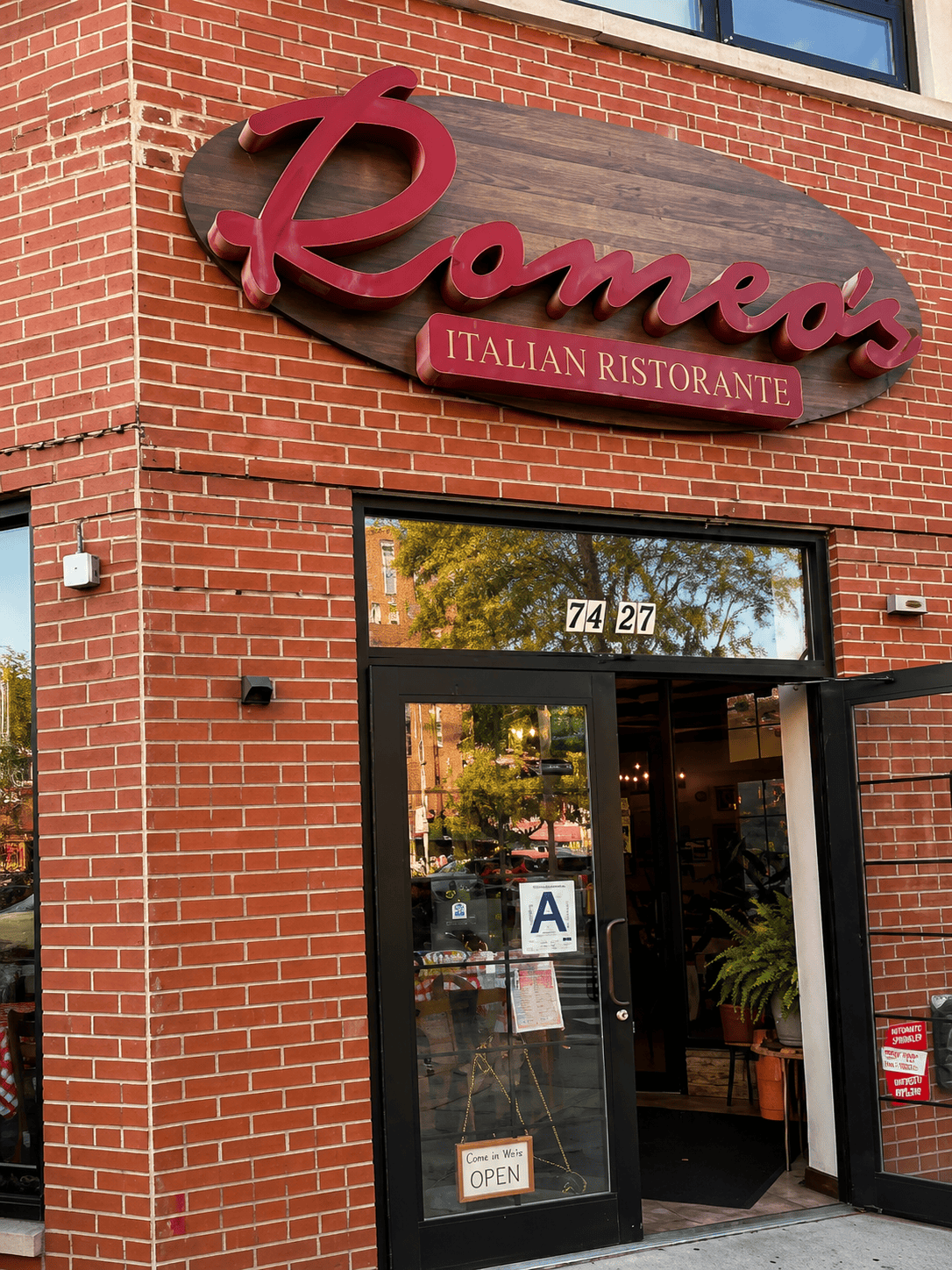 Exterior storefront of Romeo's Italian Ristorante in Jackson Heights, featuring a large oval wooden sign with red cursive text on a classic red brick building.