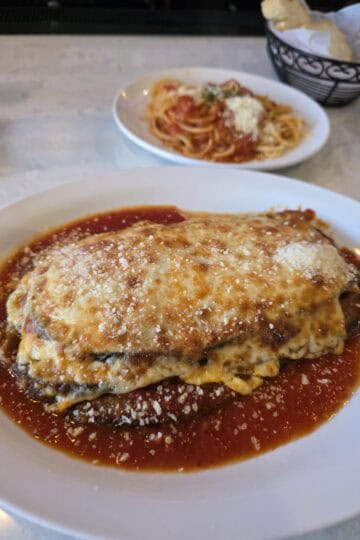 A large oval plate holding a thick portion of baked Eggplant Parmesan covered in a layer of melted mozzarella and tomato sauce, with a side plate of spaghetti in the background.