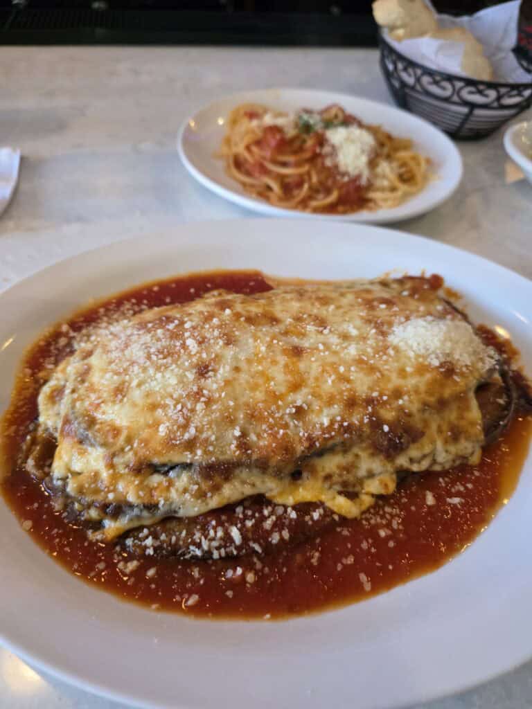 A large oval plate holding a thick portion of baked Eggplant Parmesan covered in a layer of melted mozzarella and tomato sauce, with a side plate of spaghetti in the background.