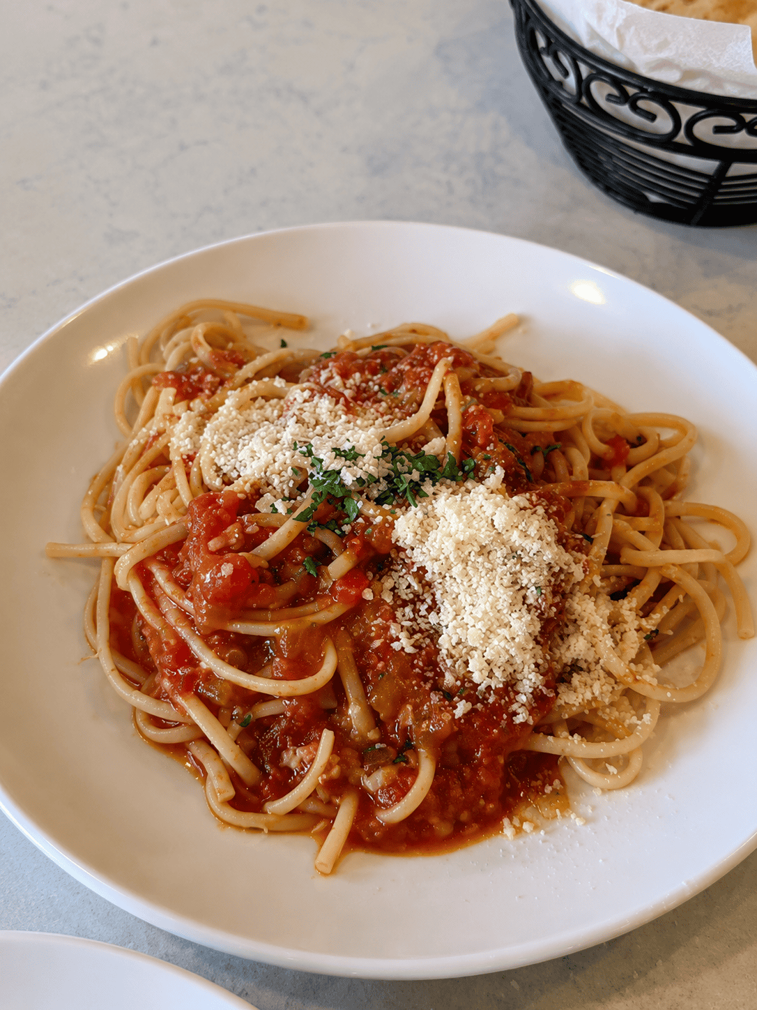 A white plate holding a side portion of spaghetti pasta tossed in a bright red tomato sauce and topped with grated Parmesan cheese and fresh parsley.