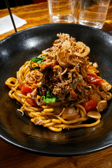 A black ceramic bowl filled with thick stir-fried noodles topped with tender shredded slow-cooked beef brisket and crispy fried shallots.