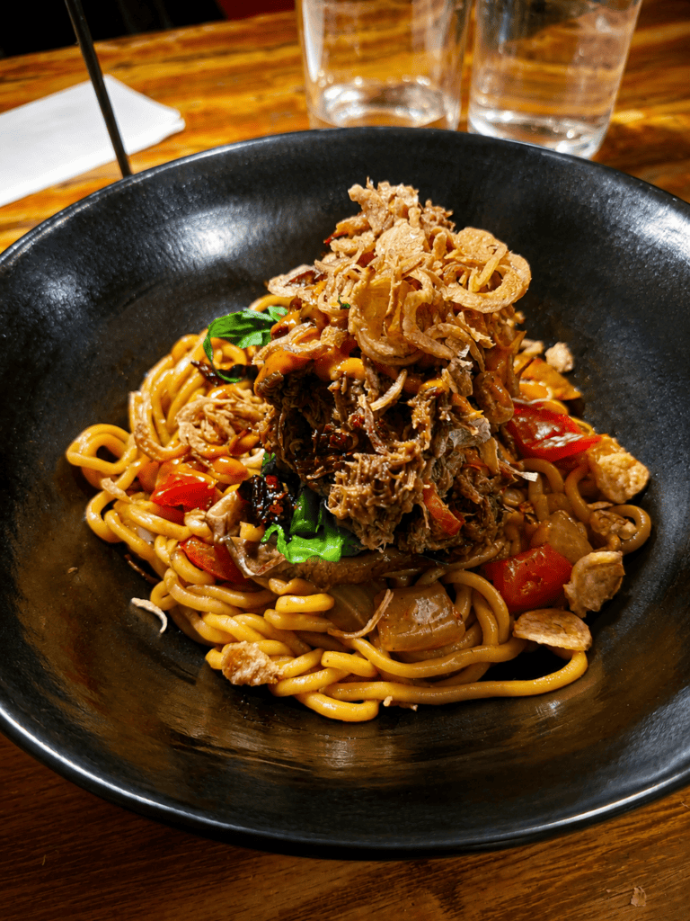 A black ceramic bowl filled with thick stir-fried noodles topped with tender shredded slow-cooked beef brisket and crispy fried shallots.