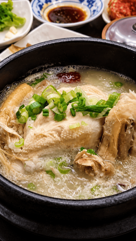 Samgyetang traditional Korean ginseng chicken soup featuring a whole young chicken stuffed with rice simmering in a bubbling broth with fresh green scallions and a jujube fruit.