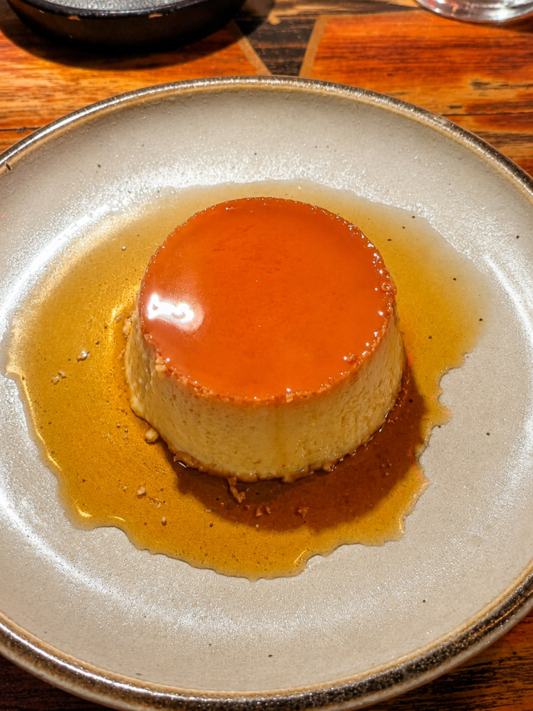 A perfectly smooth and round traditional flan custard swimming in a pool of golden caramel syrup on a rustic ceramic plate.