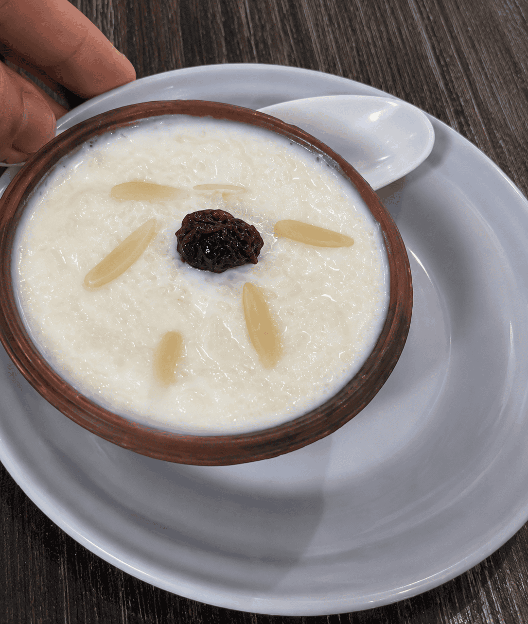 A traditional bowl of white rice pudding served in a rustic clay dish on a white saucer, garnished with almond slivers and a single dark raisin in the center.