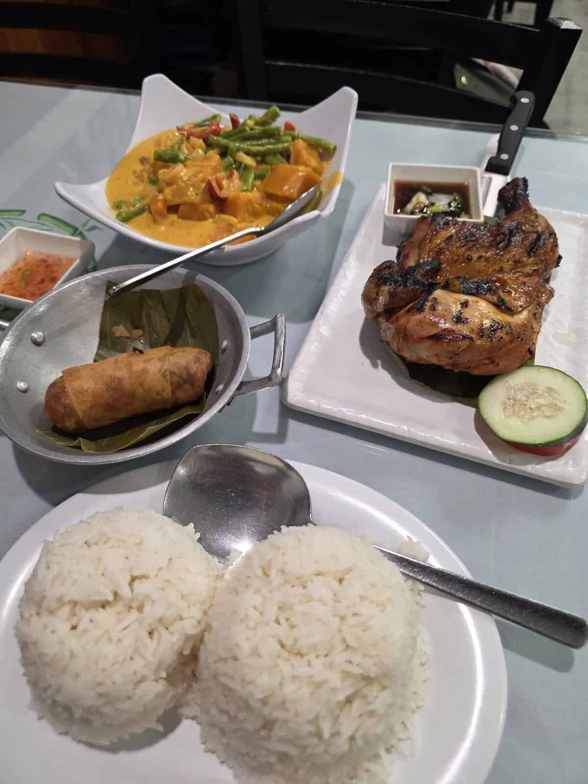 A table spread of Filipino dishes at Tyang Gelay's Grill, featuring a bowl of creamy yellow squash and green bean curry, grilled chicken inasal on a banana leaf, a fried lumpia roll, and two mounds of white rice.
