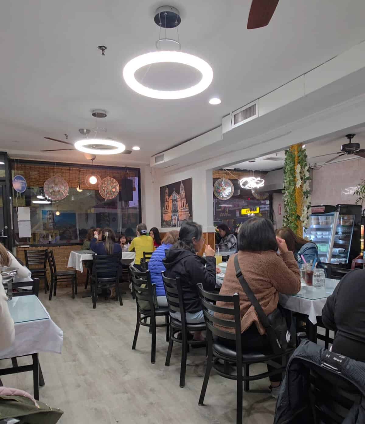 Cozy and casual interior of Tyang Gelay's Grill dining room in Woodside, filled with families and friends eating at tables under circular light fixtures.