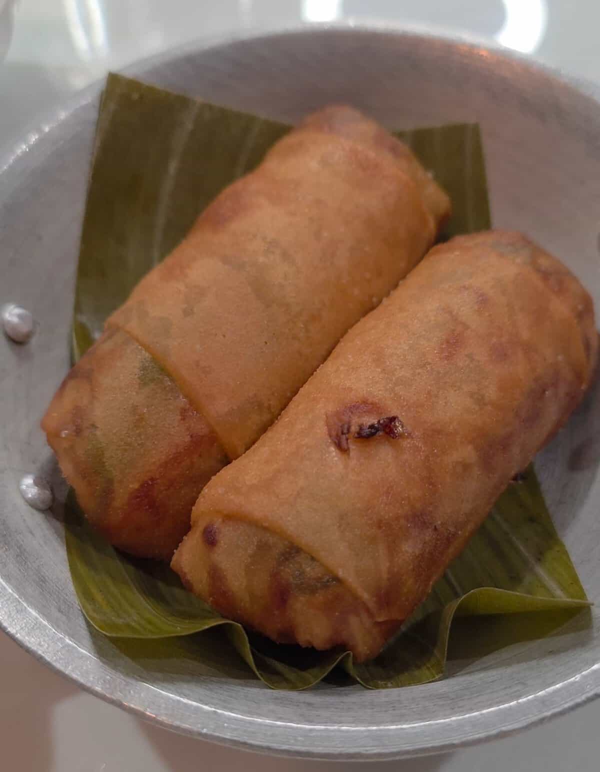Two golden-brown, crispy fried Filipino lumpia prito served on a green banana leaf inside a small silver metal dish.