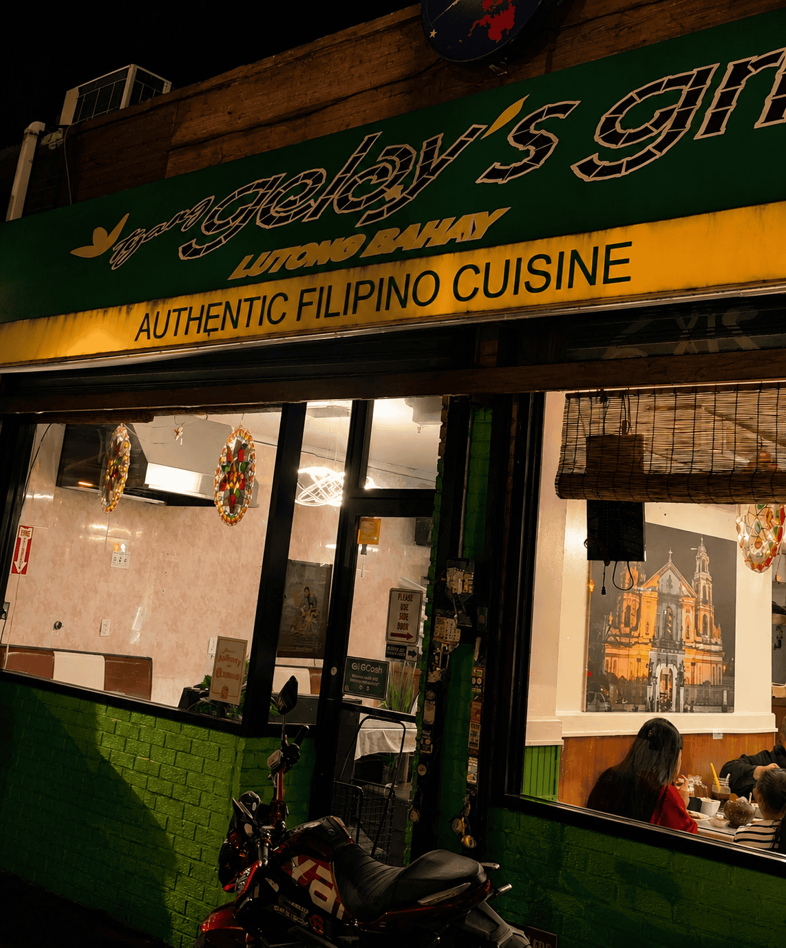 Storefront of Tyang Gelay's Grill in Woodside, Queens at night, featuring a green and yellow sign that reads "Authentic Filipino Cuisine" with a motorcycle parked in front.
