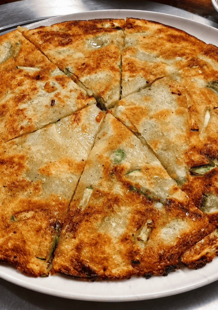 A golden brown crispy Yachae Pajun Korean vegetable pancake sliced into geometric wedges on a white plate showing scallions and zucchini ribbons.