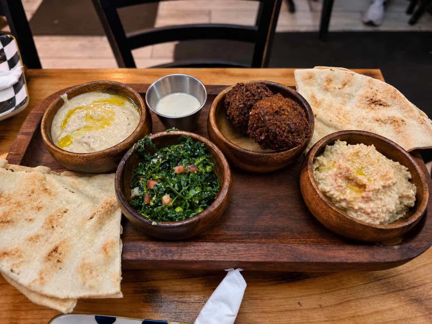 Wooden tray holding bowls of falafel tabouli hummus baba ganoush tahini sauce and pita bread