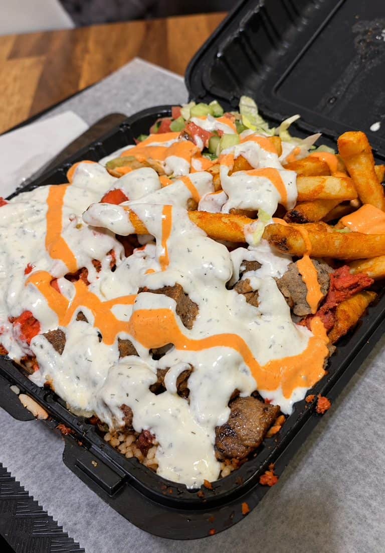 Overloaded takeout tray with chicken, lamb, spiced rice, fries, salad, white sauce, and red spicy sauce