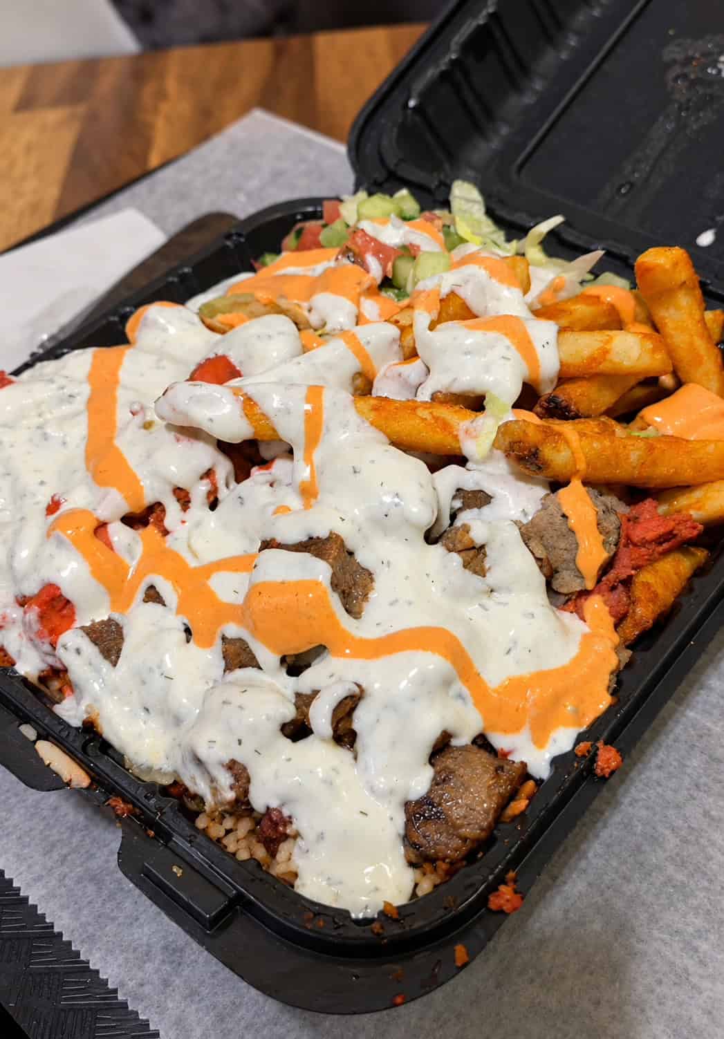 Overloaded takeout tray with chicken, lamb, spiced rice, fries, salad, white sauce, and red spicy sauce