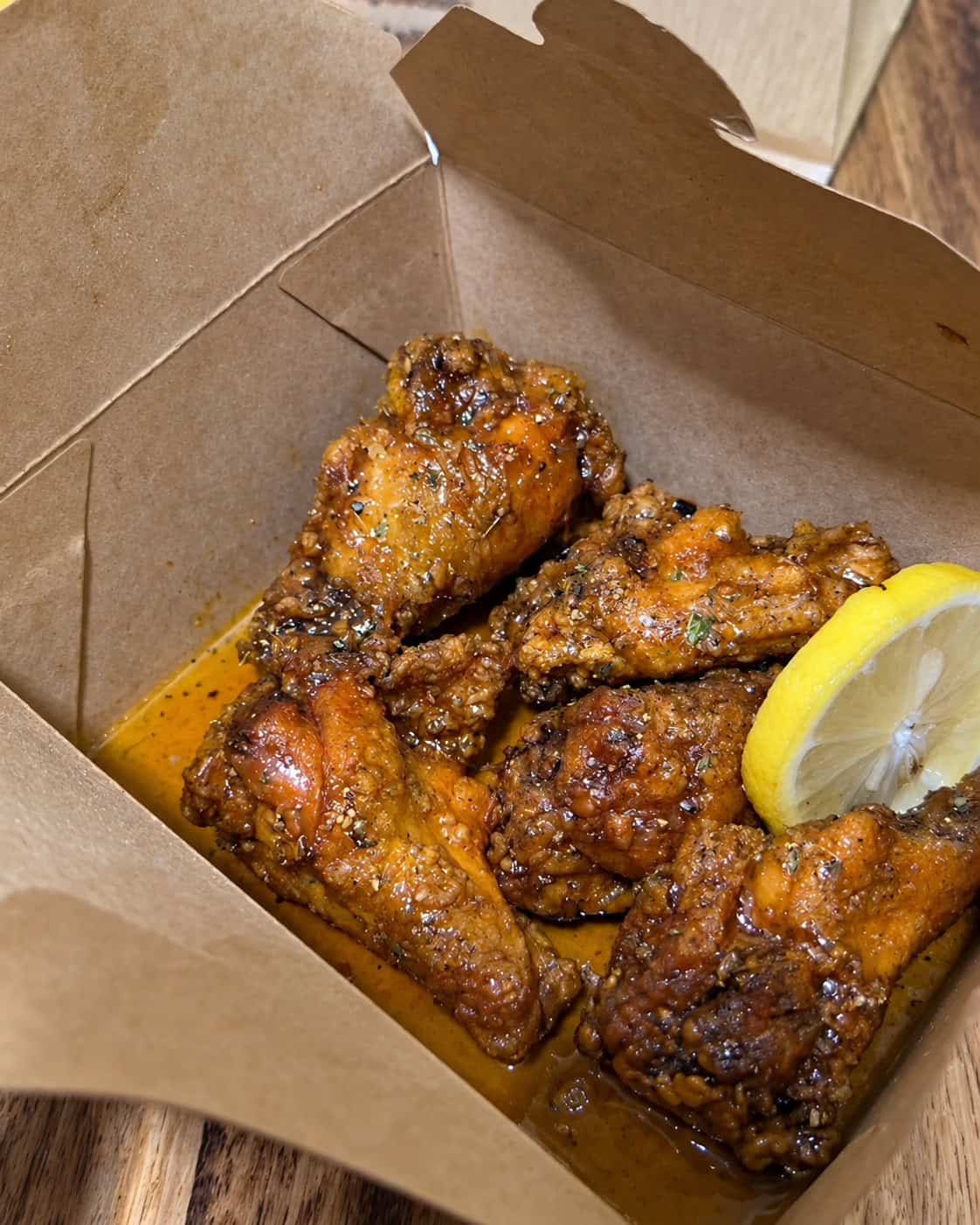 Golden fried chicken wings tossed in glazed sauce with fresh lemon wedge inside takeout box