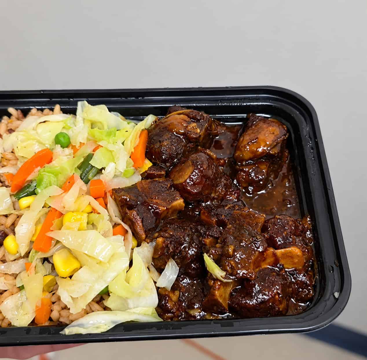 Takeout container filled with tender oxtail stew, rice and peas, and steamed cabbage