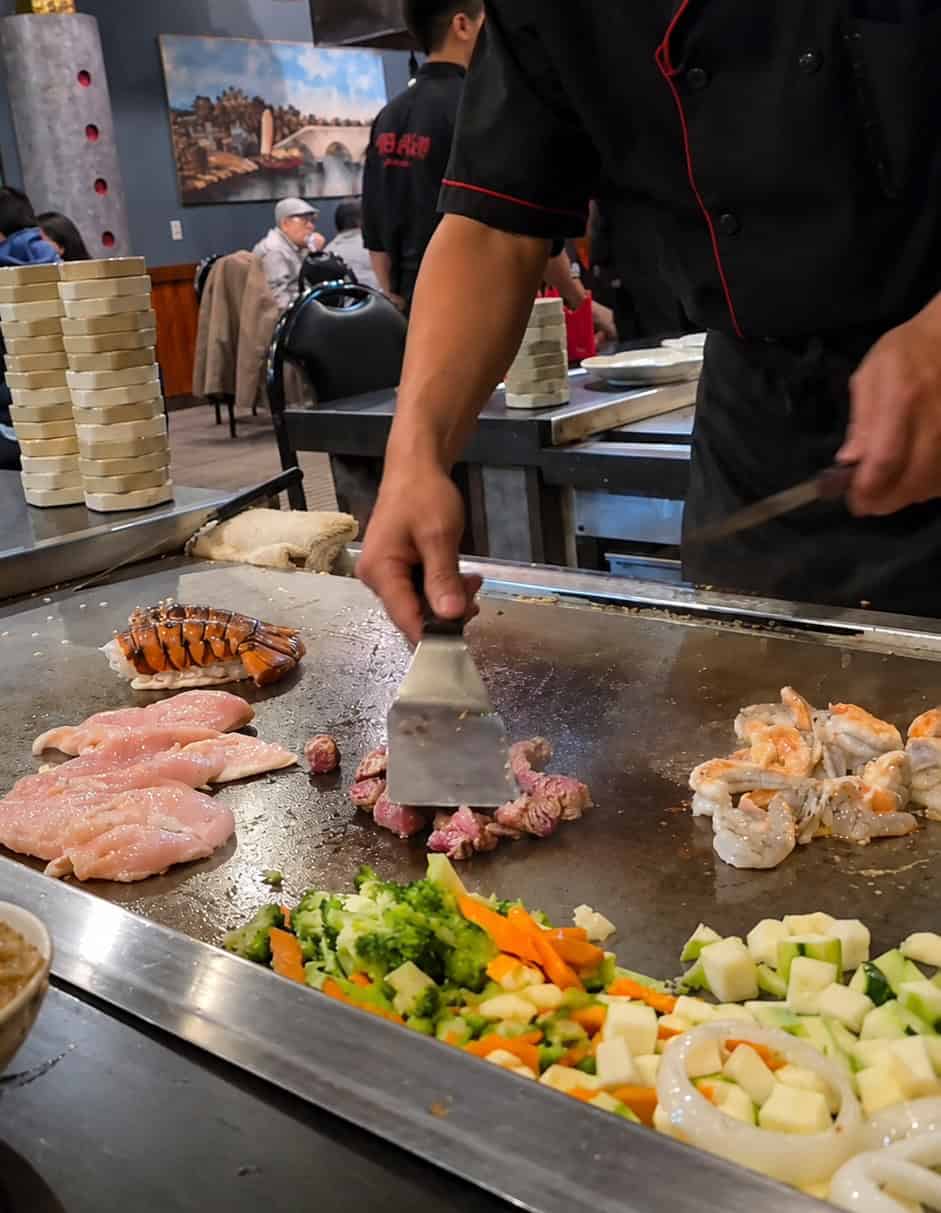 Hibachi chef using spatulas to cook chicken breast, steak cubes, shrimp, lobster tail, and chopped vegetables on the grill