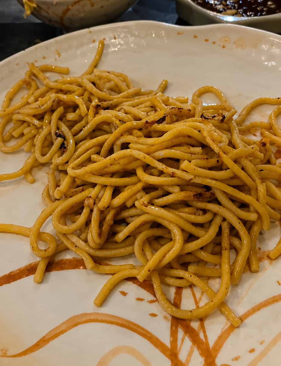 Warm plate of hibachi stir-fried lo mein noodles served with fried rice and dipping sauce in the background